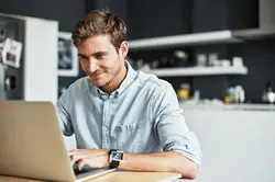 Happy young man using his laptop while sitting in his kitchen