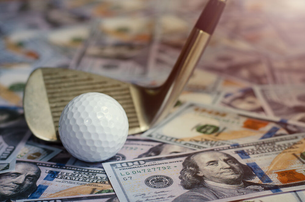 A golf ball and club rest on a messy stack of $100 bills