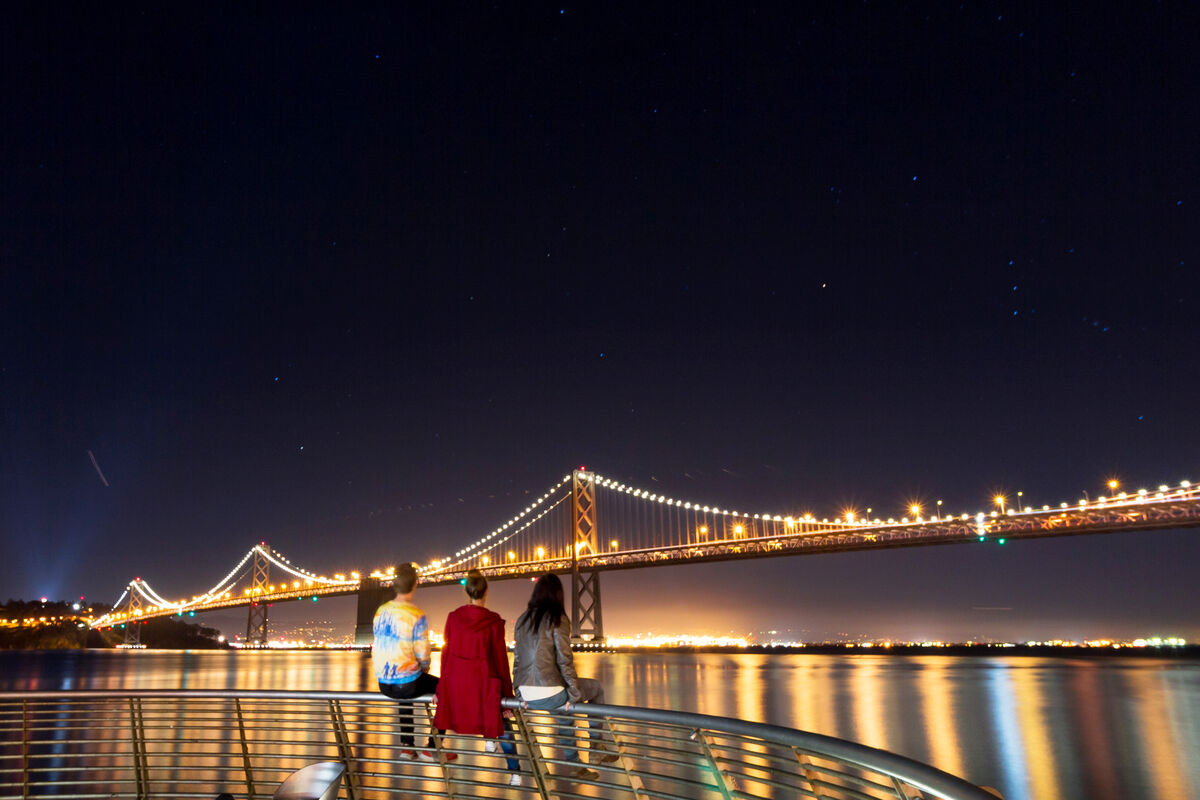 People Sitting On Railing Against Illuminated Bay Bridge At Night